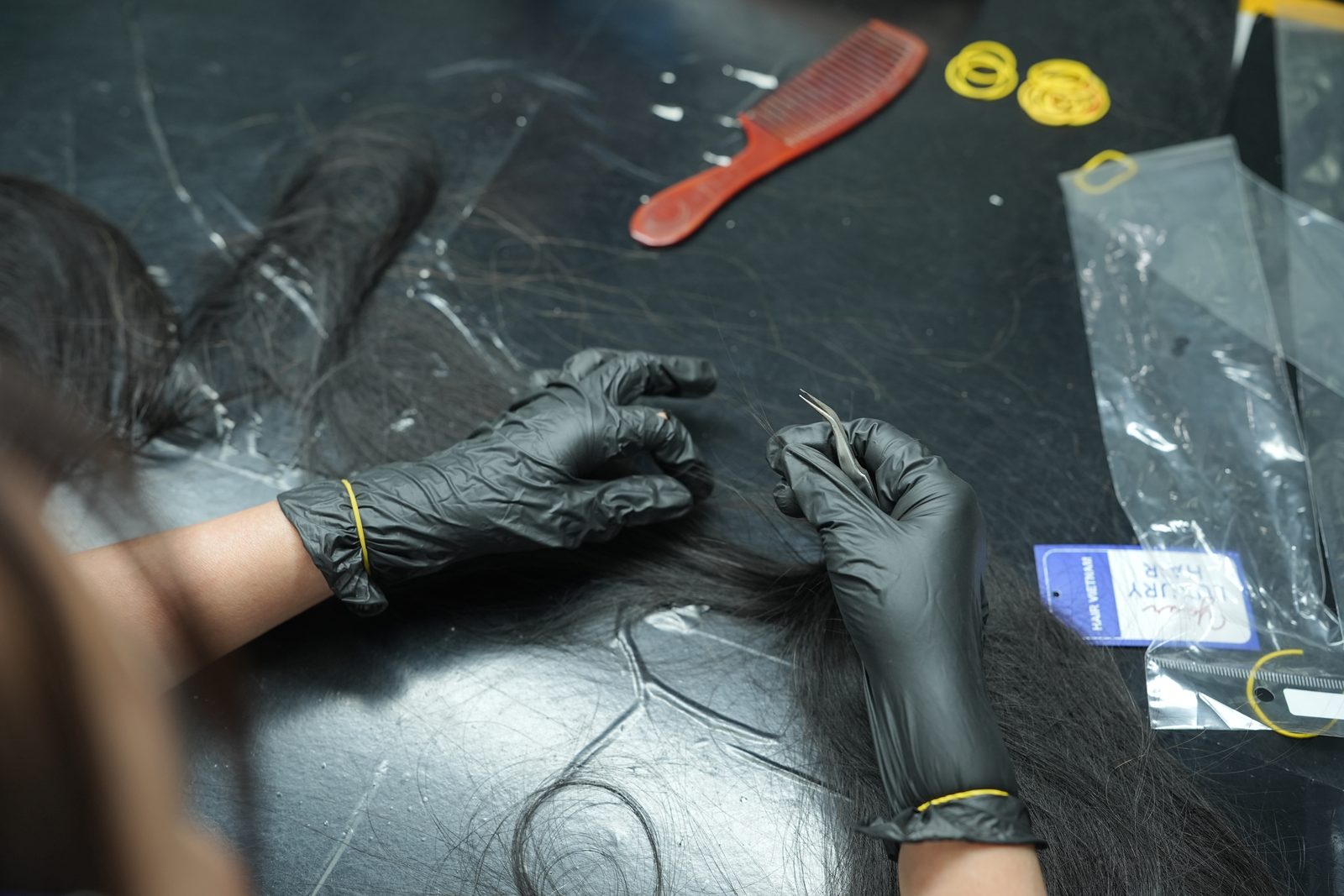 Gloved hands pick through dark hair on a black table next to a red comb and rubber bands