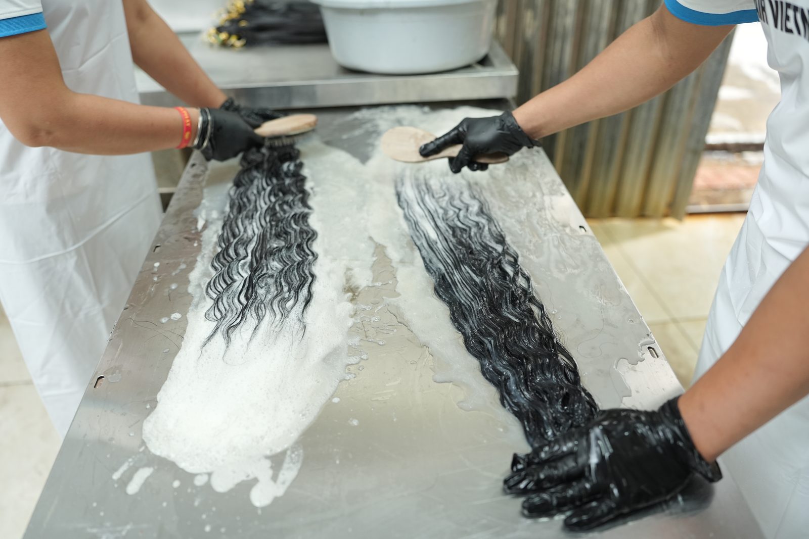 Gloved hands brush two wavy dark hair bundles covered in white foam on a stainless-steel table