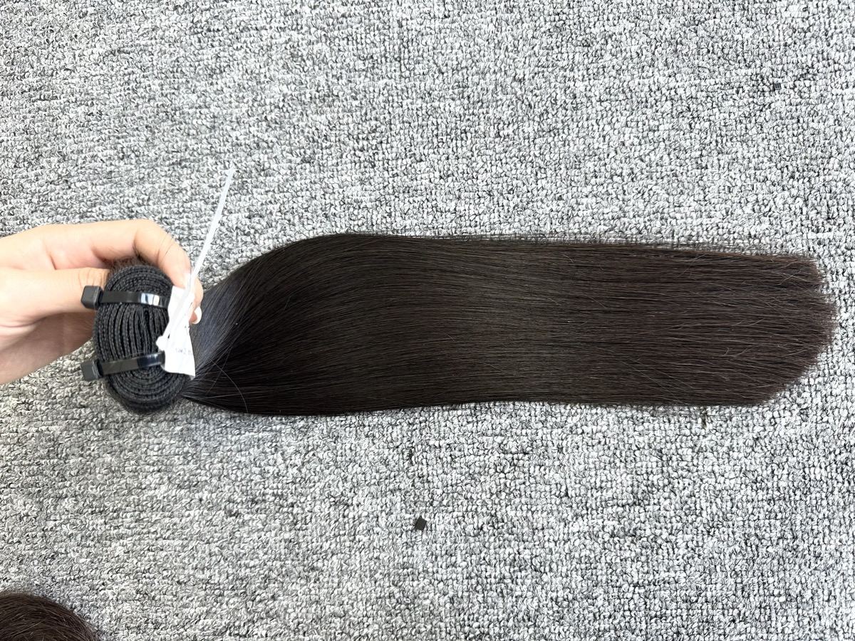 A hand holds the weft of a long straight dark hair bundle laid flat on a carpet