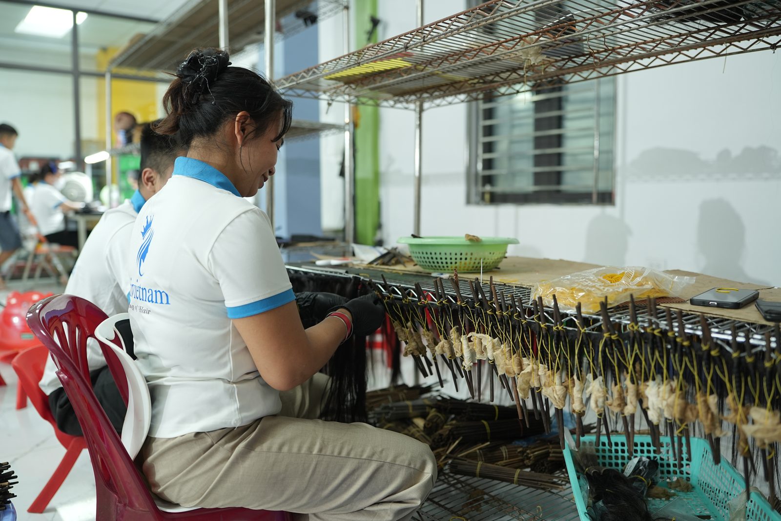 A staff member in a white polo shirt hand-works dark hair strands tied to small pegs on a wire rack