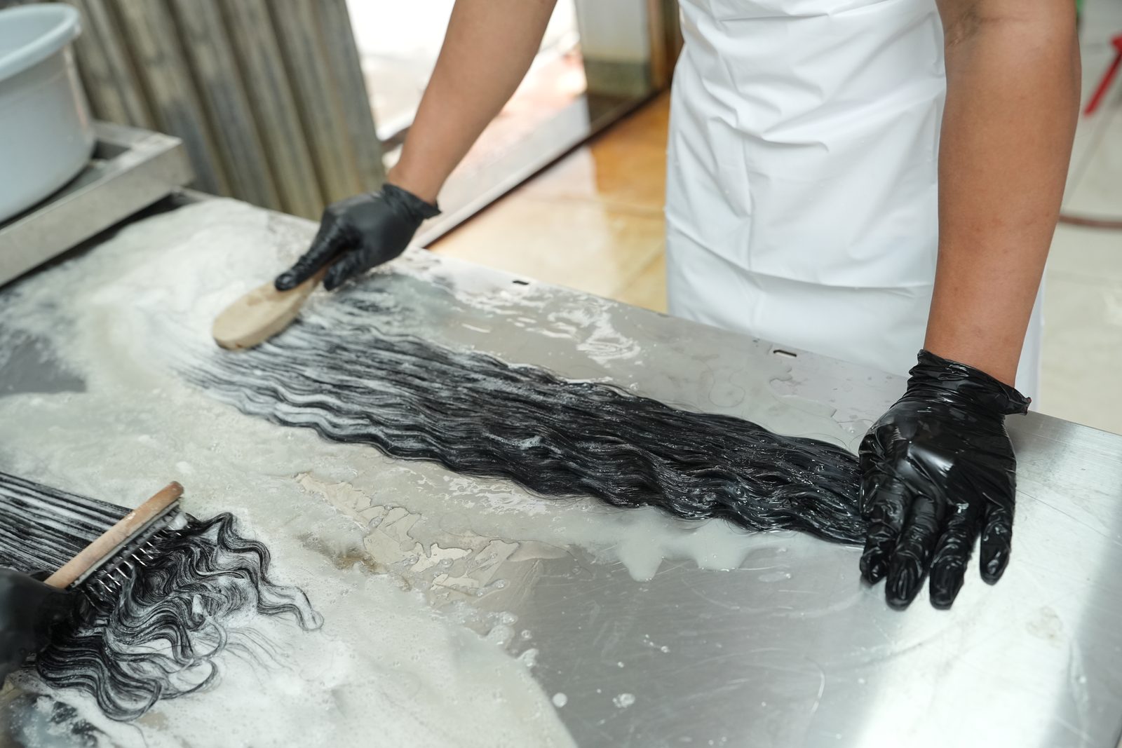 A gloved hand works a brush through a wavy dark hair bundle covered in white foam on a steel table