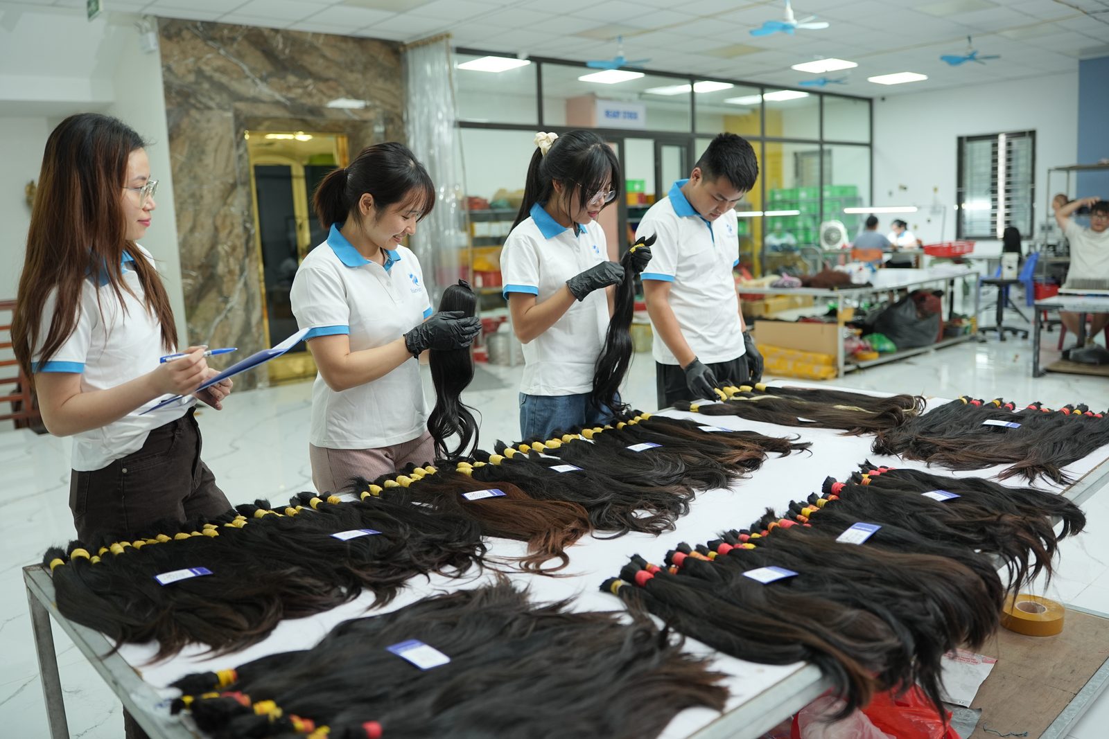 Staff inspect rows of dark hair bundles with blue tags on a long table, one holding a clipboard