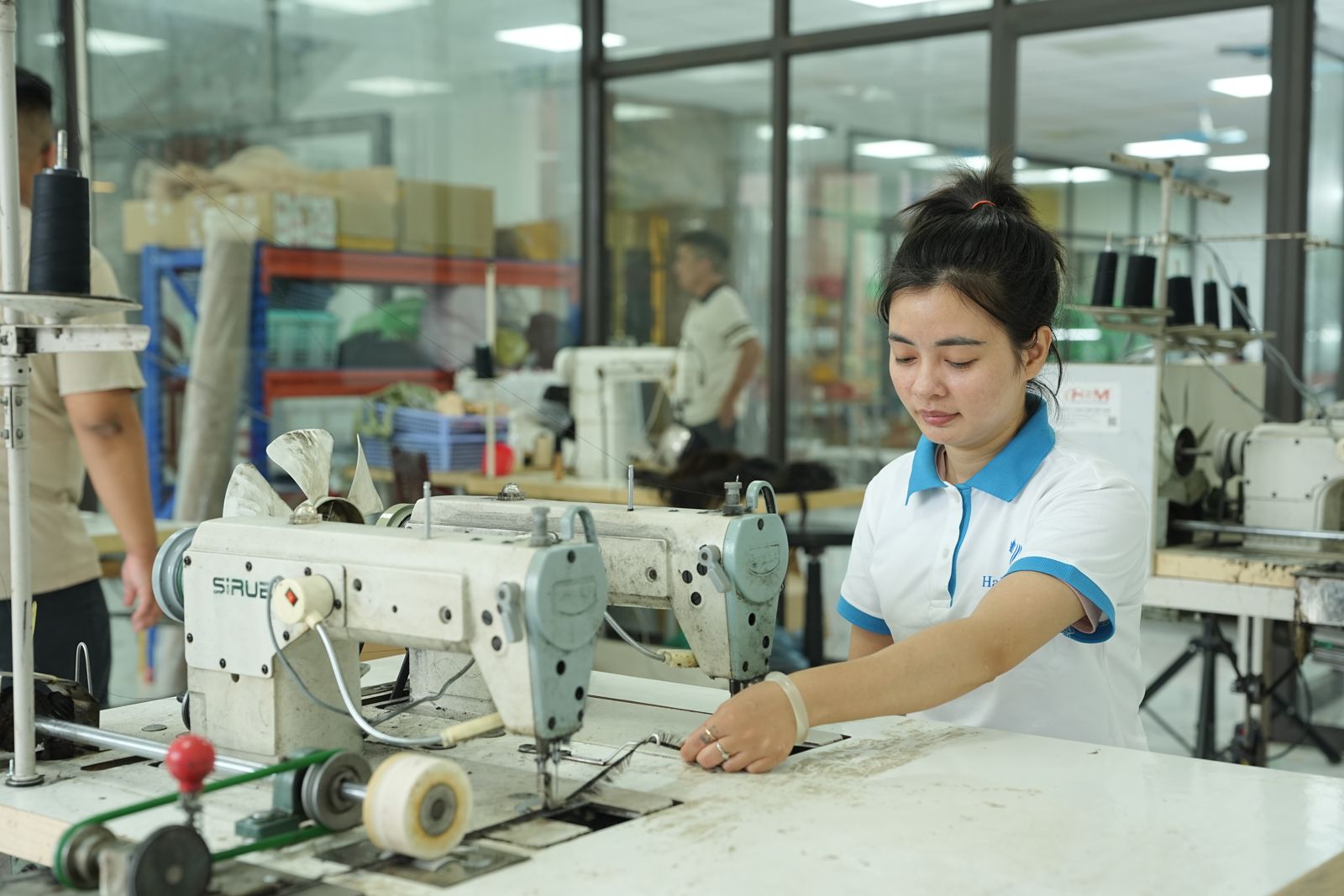 A woman in a white polo shirt sits at an industrial sewing machine in a factory workshop